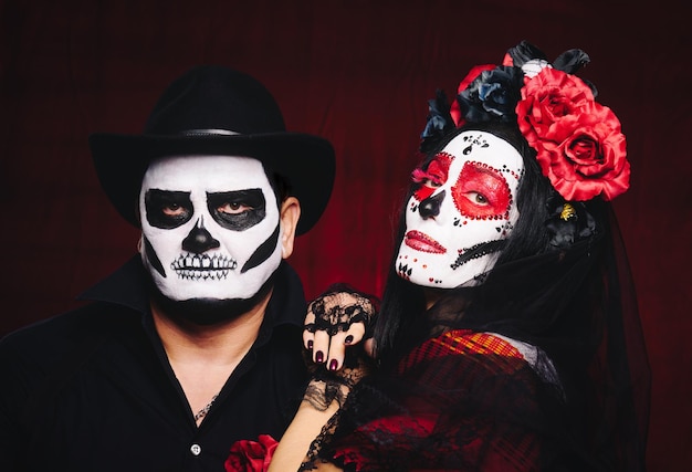 Mulher bonita com uma maquiagem de caveira de açúcar com uma coroa de flores na cabeça e um homem esqueleto com um chapéu preto