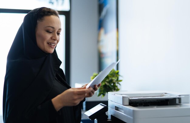 Mulher bonita com vestido abaya trabalhando e imprimindo documentos. funcionária de meia idade no trabalho em um escritório de negócios em dubai. conceito sobre culturas e estilo de vida do oriente médio