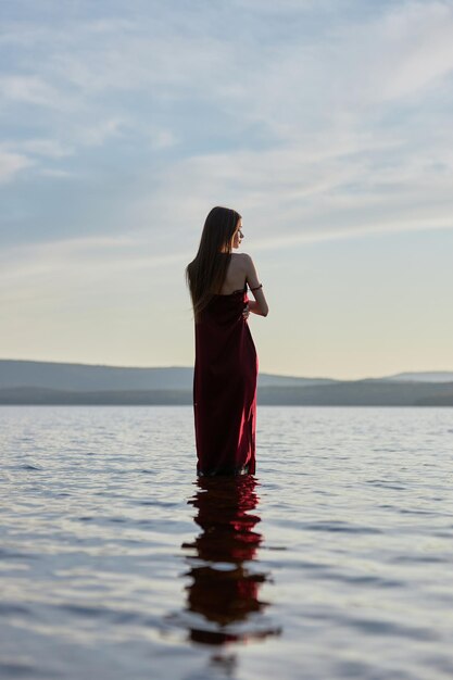 Mulher bonita com vestido vermelho claro fica na água do mar do Lago ao pôr do sol. Reflexo da menina e do céu na água