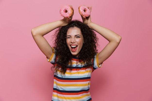 Mulher bonita de 20 anos com cabelo encaracolado se divertindo e segurando rosquinhas isoladas em rosa