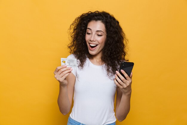 Mulher bonita de 20 anos vestindo roupas casuais segurando um telefone celular e um cartão de crédito isolado no amarelo