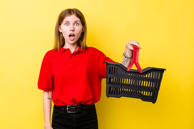 Mulher bonita de cabeça vermelha olhando muito chocado ou surpreso conceito de cesta de compras