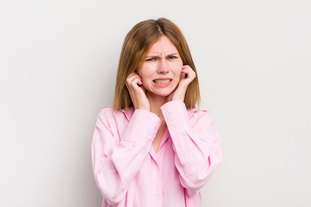 Foto mulher bonita de cabeça vermelha parecendo irritada estressada e irritada cobrindo os dois ouvidos com um som ensurdecedor ou música alta