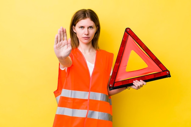 Foto mulher bonita de cabeça vermelha parecendo séria mostrando a palma aberta fazendo gesto de parada carro triângulo perigo