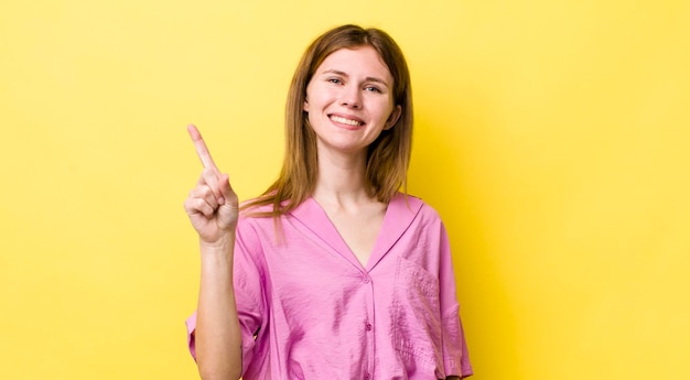 Mulher bonita de cabeça vermelha sorrindo e parecendo amigável, mostrando o número um ou primeiro com a mão em contagem regressiva