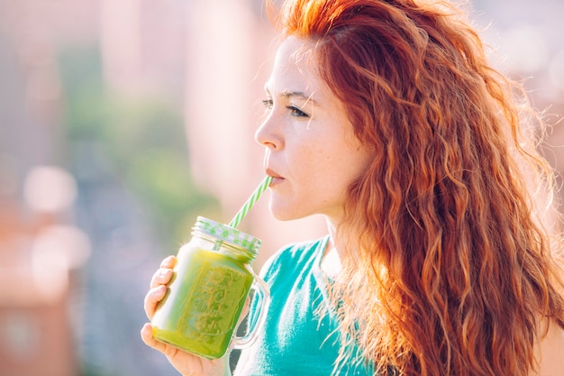 Foto mulher bonita de cabelo vermelho, bebendo com um canudo, um suco verde saudável