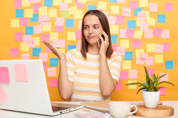 Mulher bonita de cabelos castanhos confusa e desamparada sentada à mesa em seu local de trabalho com laptop contra parede amarela falando ao telefone e trabalhando no notebook não sabe o que fazer com seu trabalho encolhendo os ombros