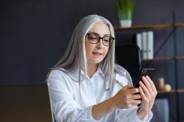 Mulher bonita de cabelos grisalhos sênior usando smartphone e sorrindo. Mulher de negócios feliz usando aplicativos de telefone celular, mensagens de texto, navegação na internet, compras online. Pessoas maduras com dispositivos móveis.