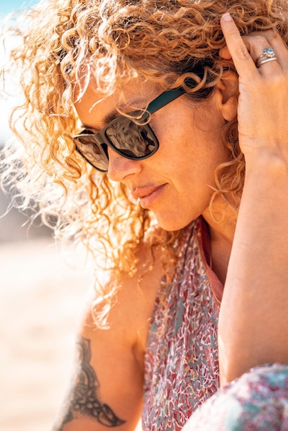 Mulher bonita de meia idade com cabelos cacheados e óculos de sol em retrato lateral ao ar livre Pessoas femininas bonitas e modernas com tatuagem relaxam na praia sob o sol de verão Vida de pessoas loiras saudáveis