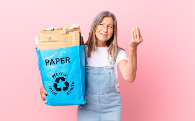 Mulher bonita de meia-idade fazendo um gesto de capice ou dinheiro, dizendo para você pagar o conceito de reciclagem de papelão