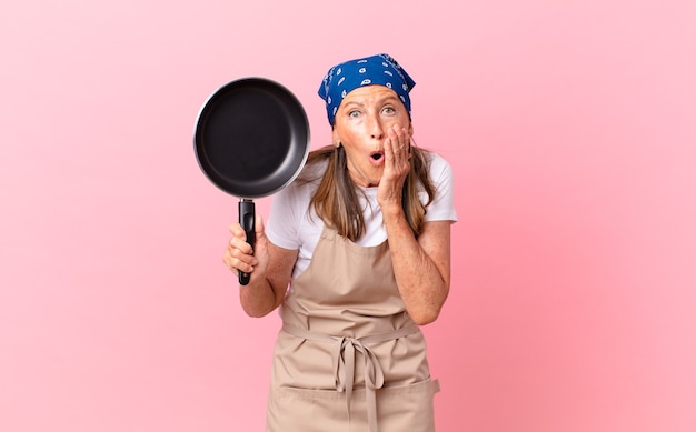 Mulher bonita de meia-idade se sentindo chocada e com medo e segurando uma panela. conceito de chef