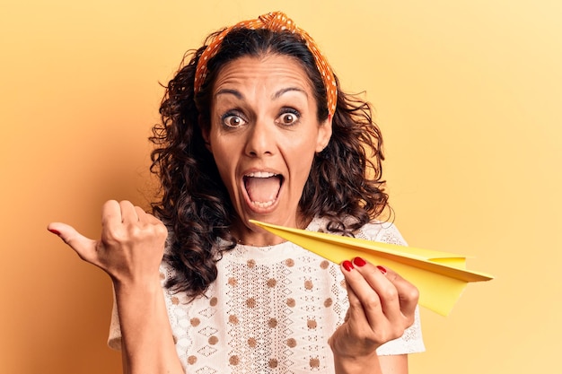 Mulher bonita de meia-idade segurando avião papar apontando o polegar para o lado sorrindo feliz com a boca aberta