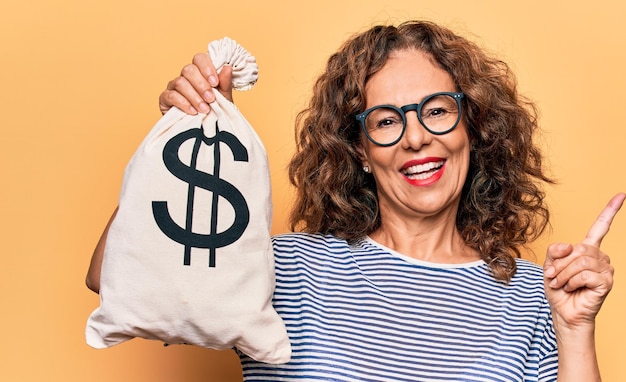 Mulher bonita de meia-idade segurando saco com símbolo de dólar sobre fundo amarelo isolado sorrindo feliz apontando com a mão e o dedo para o lado