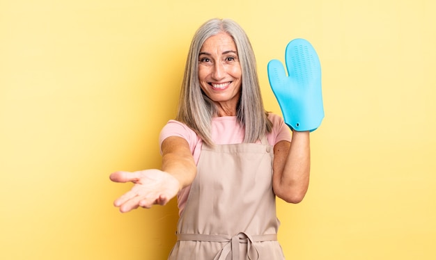 Mulher bonita de meia-idade, sorrindo alegremente com simpáticos e oferecendo e mostrando um conceito. conceito de luva de forno