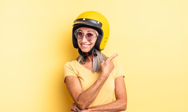 Mulher bonita de meia-idade sorrindo alegremente, se sentindo feliz e apontando para o lado. conceito de capacete de motocicleta