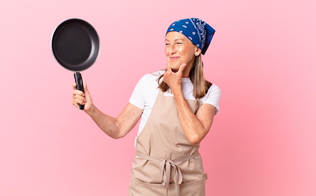 Mulher bonita de meia-idade, sorrindo com uma expressão feliz e confiante, com a mão no queixo e segurando uma panela. conceito de chef