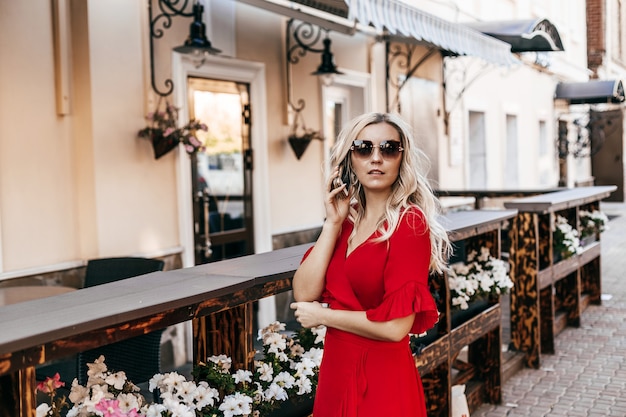 Mulher bonita de óculos escuros e vestido vermelho falando ao telefone perto de um café de rua