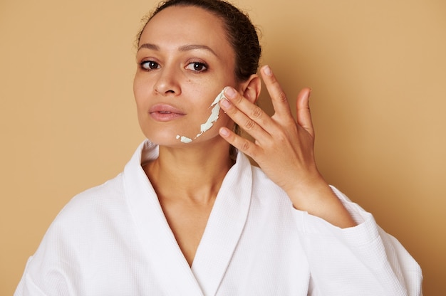 Mulher bonita de raça mista, aplicando uma máscara de beleza no rosto. retrato tirado na cabeça em superfície bege com espaço de cópia