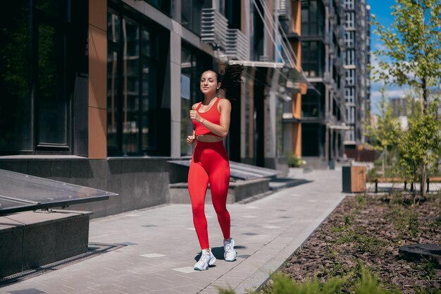 Mulher bonita desportiva correndo na calçada de azulejos na área urbana. Menina apta com rabo de cavalo escuro, vestindo um top vermelho esporte, leggins e sheakers brancos. Bloco de apartamentos e árvores no fundo.