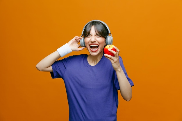 Mulher bonita desportiva em roupas esportivas com fones de ouvido na cabeça segurando uma maçã olhando para a câmera feliz e animada sorrindo amplamente em pé sobre fundo laranja