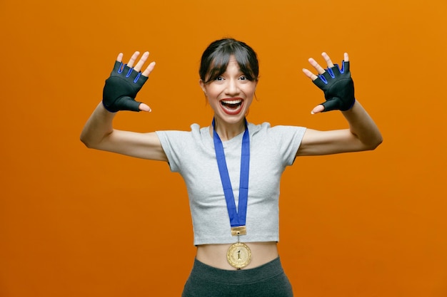 Mulher bonita desportiva em roupas esportivas em luvas com medalha no pescoço, olhando para a câmera feliz e animada, regozijando-se com seu sucesso com os braços levantados em pé sobre fundo laranja