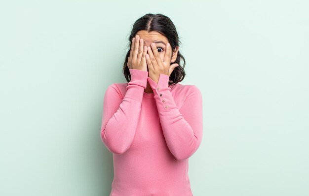 Mulher bonita e adolescente cobrindo o rosto com as mãos, espiando por entre os dedos com expressão de surpresa e olhando para o lado