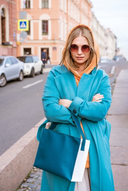 Mulher bonita e alegre com um casaco azul e um suéter laranja com uma bolsa na mão, que caminha pela cidade em um dia de primavera