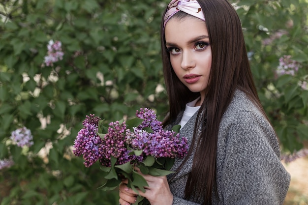 Mulher bonita e árvores floridas. Modelo de linda garota adorável no elegante casaco cinza com incríveis flores roxas é curtir a natureza no parque. Mulher atraente com buquê de lilases ao ar livre. Tempo de primavera.