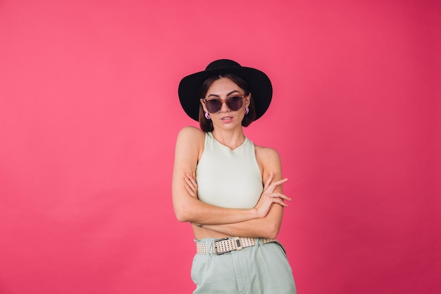 Mulher bonita e elegante com chapéu e óculos escuros posando sobre uma parede rosa vermelha