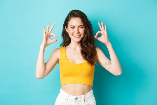 Mulher bonita e elegante, confiante, dando sinais de bom, aprovo uma boa escolha, vestindo roupas de verão, em pé sobre um fundo azul