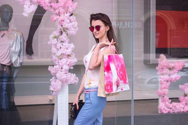 Mulher bonita e elegante de óculos fica perto das boutiques da cidade e guarda pacotes