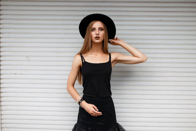 Mulher bonita e elegante em roupas da moda pretas com um chapéu perto da parede branca