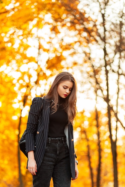 Mulher bonita e elegante em um terno preto com suéter blazer, jeans e uma mochila que caminha em um parque de outono com folhagem amarela ao pôr do sol