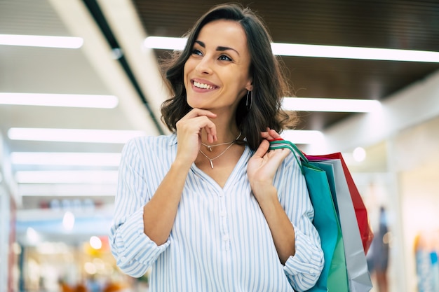 Mulher bonita e elegante jovem feliz com sacolas de compras usando o telefone inteligente enquanto caminha no shopping