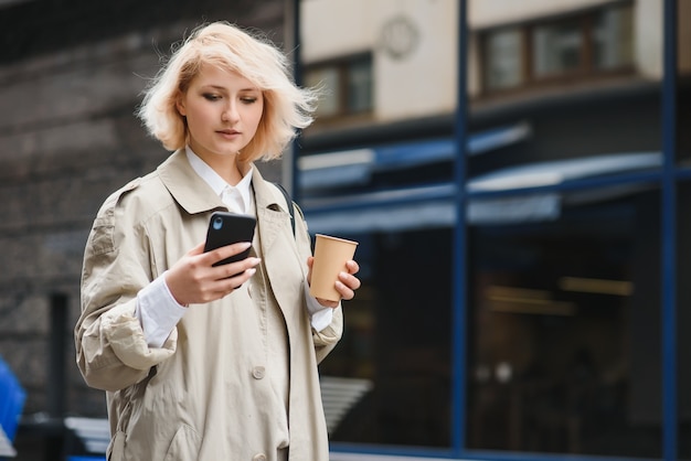 Mulher bonita e elegante tendo boas roupas da moda, andando na rua e bebendo um café na xícara para viagem com bom humor de verão, perto do café de rua.