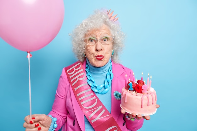 Mulher bonita e enrugada de cabelos grisalhos mantém os lábios dobrados usa maquiagem brilhante contém bolo delicioso celebra o 102º aniversário indo soprar velas e fazer desejo segura balão inflado. Conceito de hora de festa