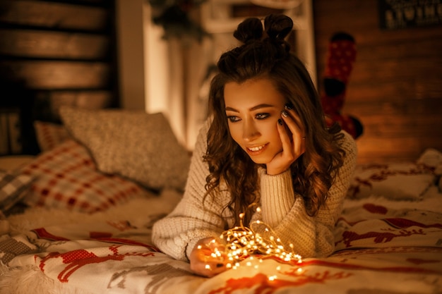 Mulher bonita e feliz em um suéter vintage com as luzes do feriado, sonhando em uma cama na véspera do natal