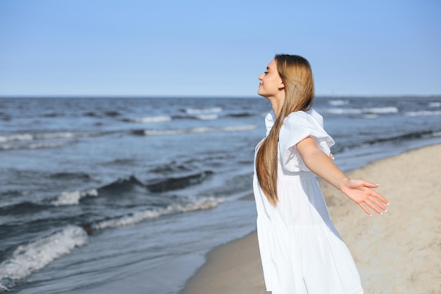 Mulher bonita e feliz na praia do oceano em pé em um vestido branco de verão, braços abertos.