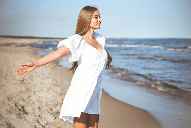 Mulher bonita e feliz na praia oceânica parada em um vestido branco de verão, braços abertos