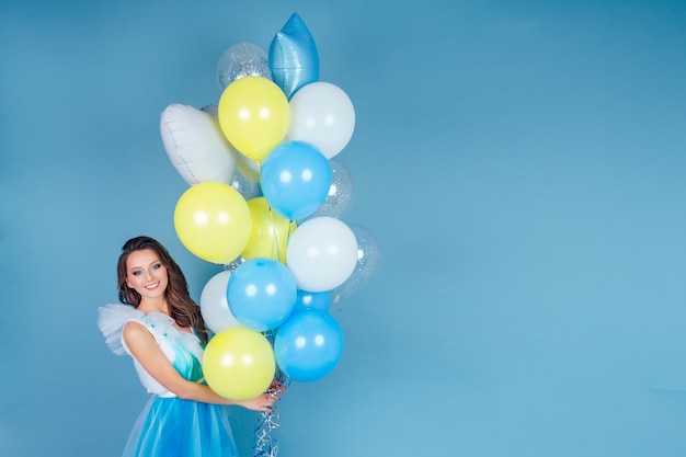 Mulher bonita e jovem com maquiagem e penteado segurando balões de hélio de ar na mão no estúdio em um fundo azul cópia space.circus e felicidade concept.prom idea