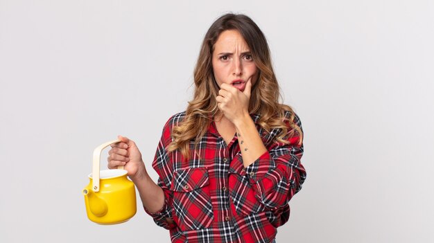 Mulher bonita e magra com boca e olhos bem abertos, mão no queixo e segurando um bule de chá