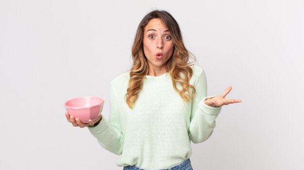 Mulher bonita e magra espantada, chocada e atônita com uma surpresa inacreditável e segurando uma tigela de panela vazia