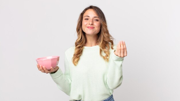 Mulher bonita e magra fazendo um gesto de capice ou dinheiro, dizendo para você pagar e segurando uma tigela vazia