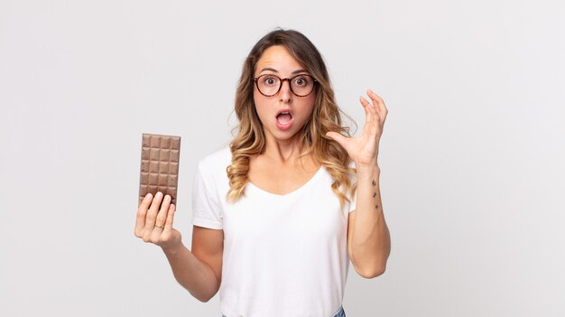 Mulher bonita e magra gritando com as mãos para o alto e segurando uma barra de chocolate