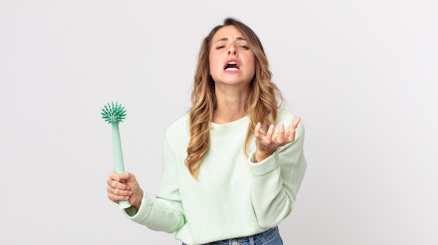 Mulher bonita e magra parecendo desesperada, frustrada e estressada segurando uma escova de lavar louça