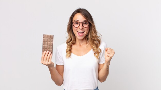 Mulher bonita e magra se sentindo chocada, rindo e comemorando o sucesso e segurando uma barra de chocolate