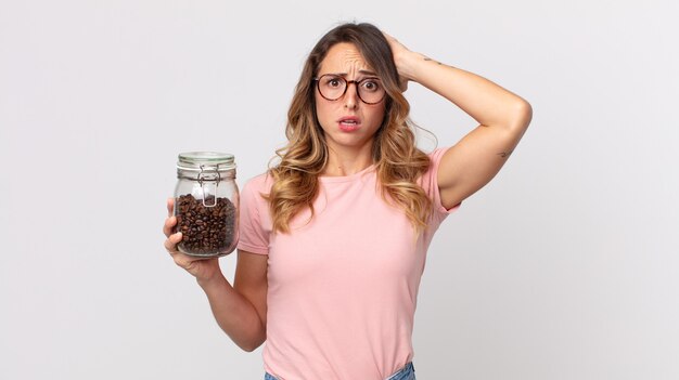 Mulher bonita e magra se sentindo estressada, ansiosa ou com medo, com as mãos na cabeça e segurando uma garrafa de grãos de café