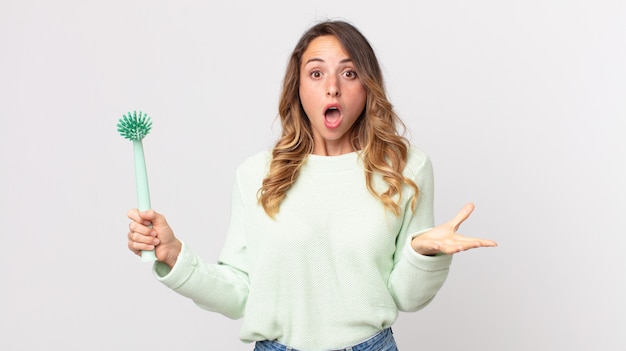 Mulher bonita e magra se sentindo extremamente chocada e surpresa, segurando uma escova de lavar louça