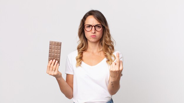 Mulher bonita e magra se sentindo irritada, irritada, rebelde e agressiva segurando uma barra de chocolate