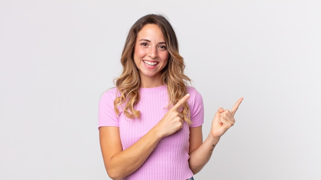 Mulher bonita e magra sorrindo alegremente e apontando para o lado e para cima com as duas mãos mostrando o objeto no espaço da cópia